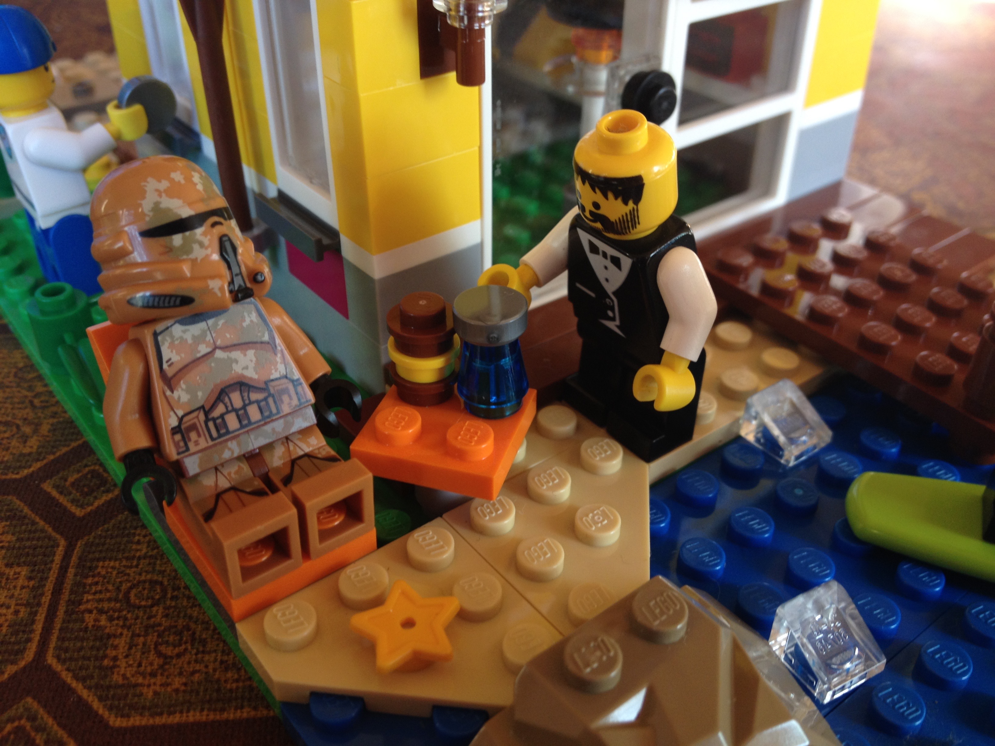 A lego waiter is giving a lego stormtrooper a lego grilled cheese sandwich and a lego cup of water, the lego stormtrooper is sitting in a lego deck chair next to the beach.