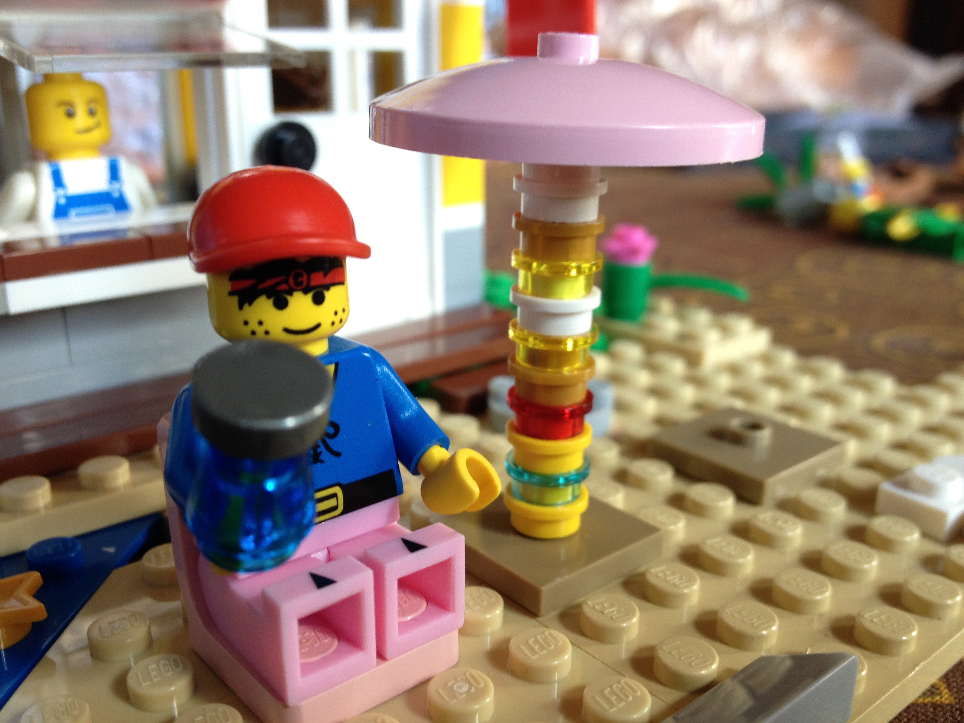 A lego person is drinking from a lego waterbottle. The lego person is sitting in a pink chair on the beach next to a striped umbrella.